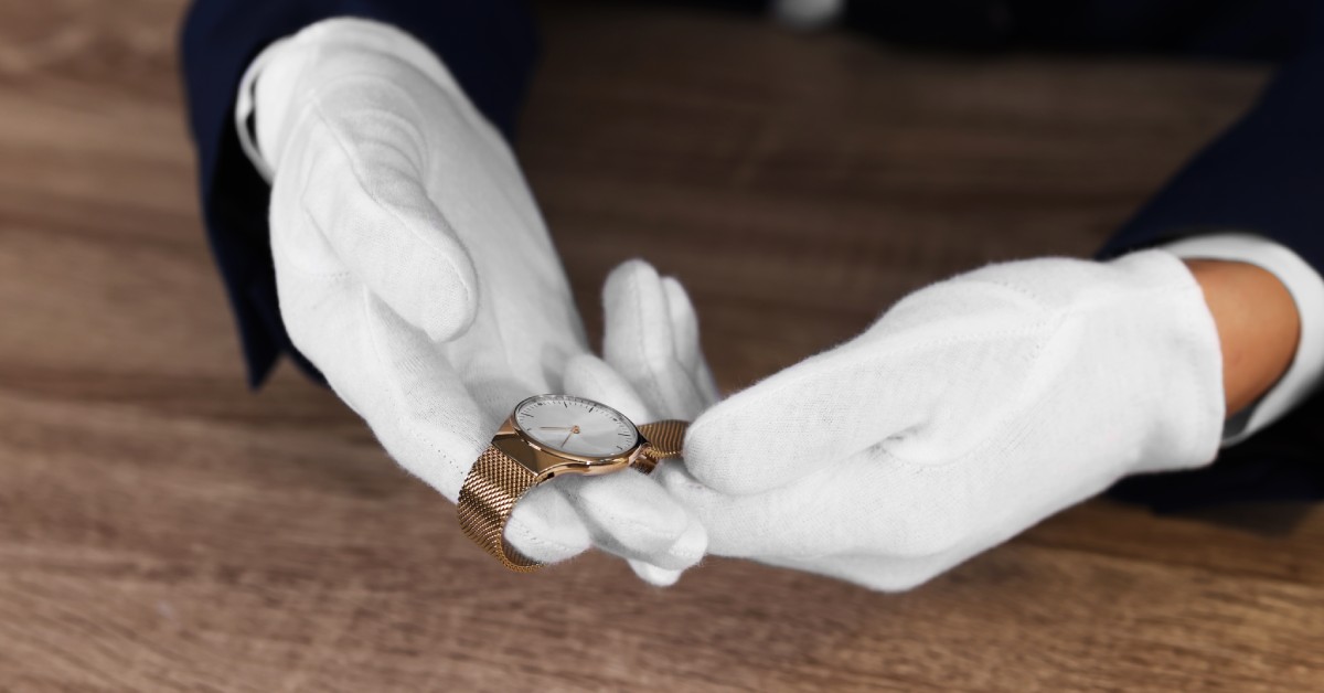 A close-up of two hands wearing white cotton gloves, holding a thin gold watch with a white face over a wooden counter.