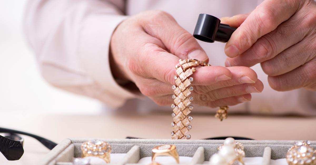 A close-up of the hands of a man holding a golden bracelet with diamonds, using a magnifying glass to look at it.